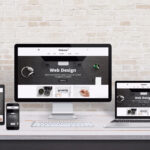 Multiple display devices with modern flat design web site presentation. Wooden desk and brick wall interior.